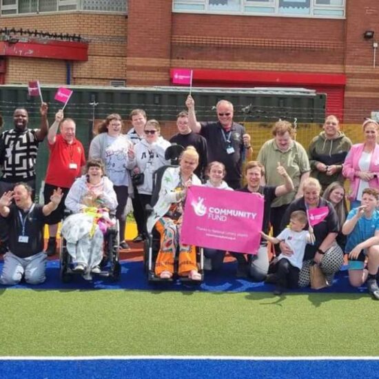 A large, diverse group of adults and children, including wheelchair users, pose together outside Daisy Inclusive UK holding pink flags and a “Community Fund” banner on a sports court in front of a brick building.