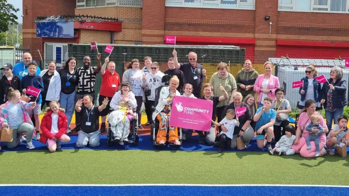 A large, diverse group of adults and children, including wheelchair users, pose together outside Daisy Inclusive UK holding pink flags and a “Community Fund” banner on a sports court in front of a brick building.
