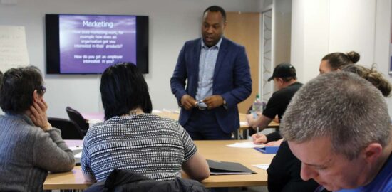 A guest presenter giving a talk to a group of Crosby Training learners. They appear to be listening intently. A man in the foreground is taking notes.