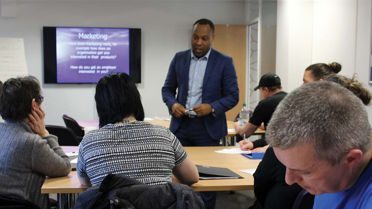 A guest presenter giving a talk to a group of Crosby Training learners. They appear to be listening intently. A man in the foreground is taking notes.