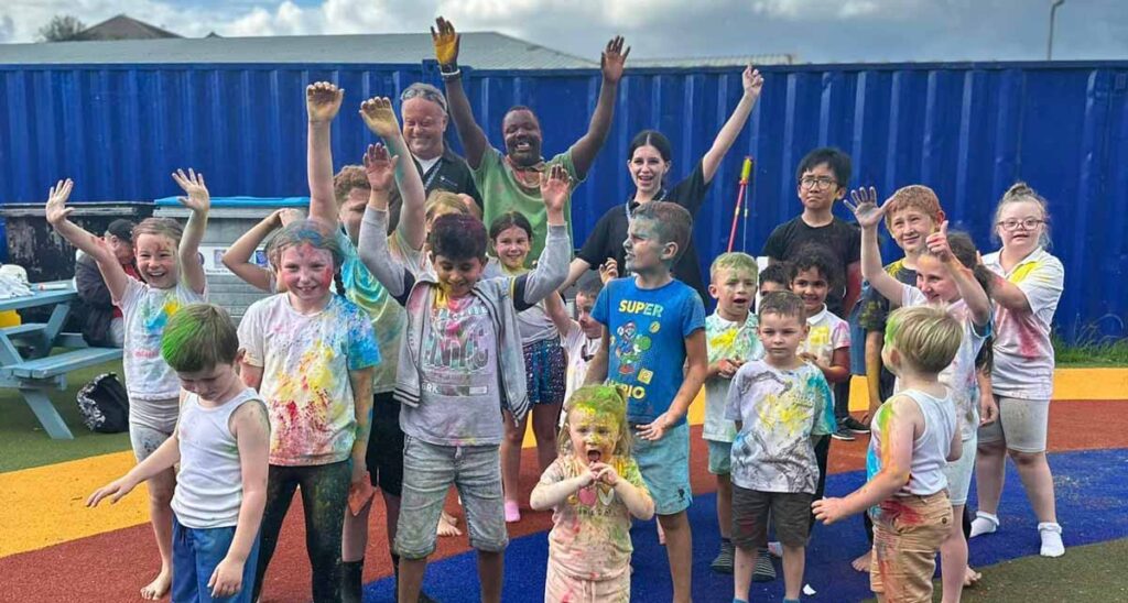 A large group of Daisy UK kids, aged between around 3 and 10, covered in various amounts of different coloured paints, looking delighted. Three equally delighted members of staff are in the background.