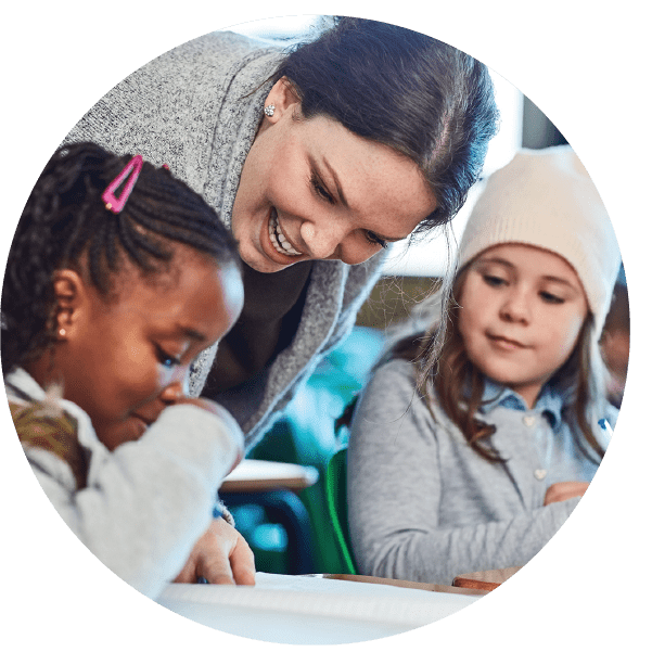 A smiling teaching assistant leaning over to help two young students with their work in a classroom