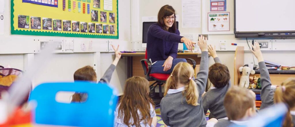 A teacher with glasses sitting down, smiling and reaching out towards primary school children sitting on the floor with their hands up in a brightly lit classroom.
