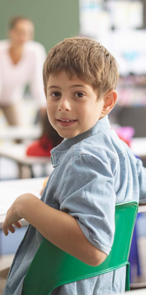 A young boy in a classroom setting, sitting on a green chair and smiling while looking back over his shoulder at the camera.