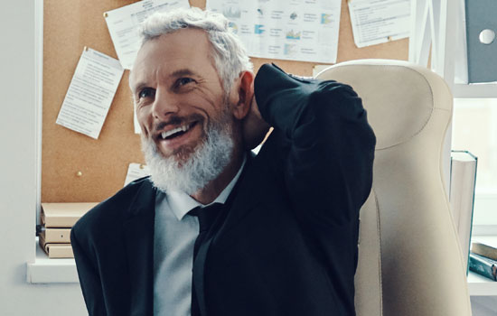 An older man in a suit and tie leaning back on his chair, hand behind his head.