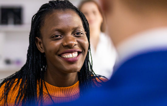 A woman smiling broadly, speaking to someone with their back to us.
