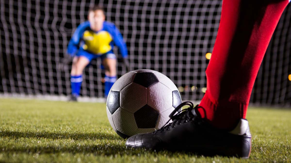 A close-up of a football on a grass pitch beside a player’s black boot and red socks, with a goalkeeper in a blue and yellow kit standing alert in front of the goal in the background.