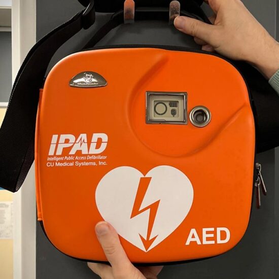 A bright orange Defibrillator hanging on a clothes peg. The livery reads IPAD AED.