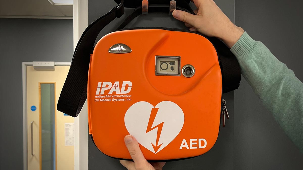 A bright orange Defibrillator hanging on a clothes peg. The livery reads IPAD AED.
