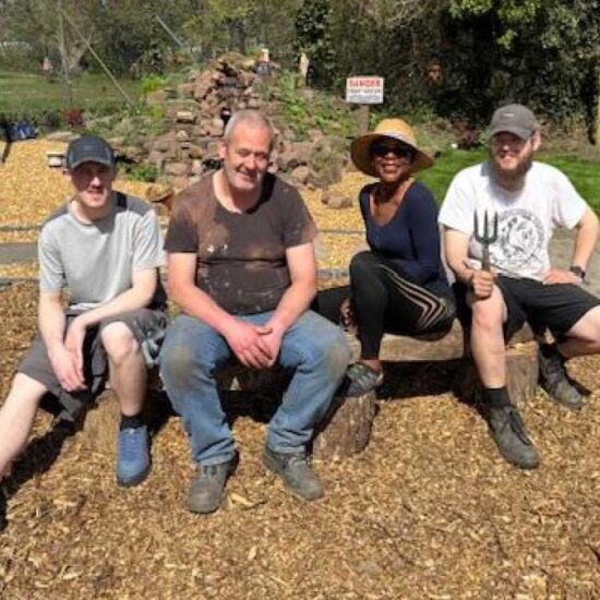 A group of four learners, three men and a woman, sitting proudly in a garden feature they have built. A gravel path with rustic flags winds past a rock formation which is the beginnings of a water feature.
