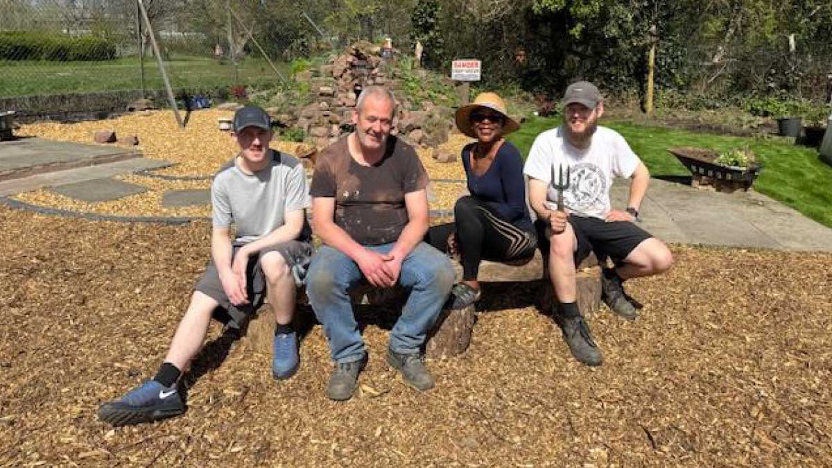 A group of four learners, three men and a woman, sitting proudly in a garden feature they have built. A gravel path with rustic flags winds past a rock formation which is the beginnings of a water feature.