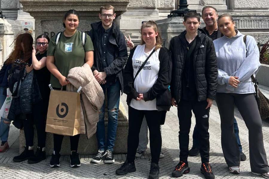 Seven young learners and their tutor outside the British Music Experience. They are dressed casually in jackets and hoodies, with one person holding a shopping bag and another holding a coat. 