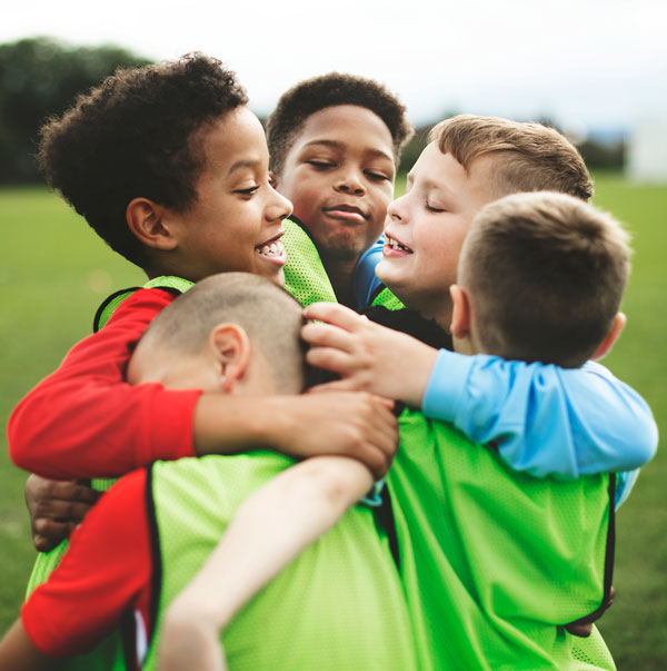 A group of young children in sports bibs hugging each other in celebration on a grassy football pitch.