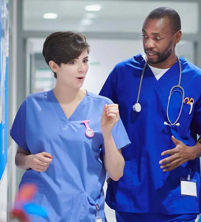 Two healthcare workers in blue scrubs walking and talking in a hospital corridor.