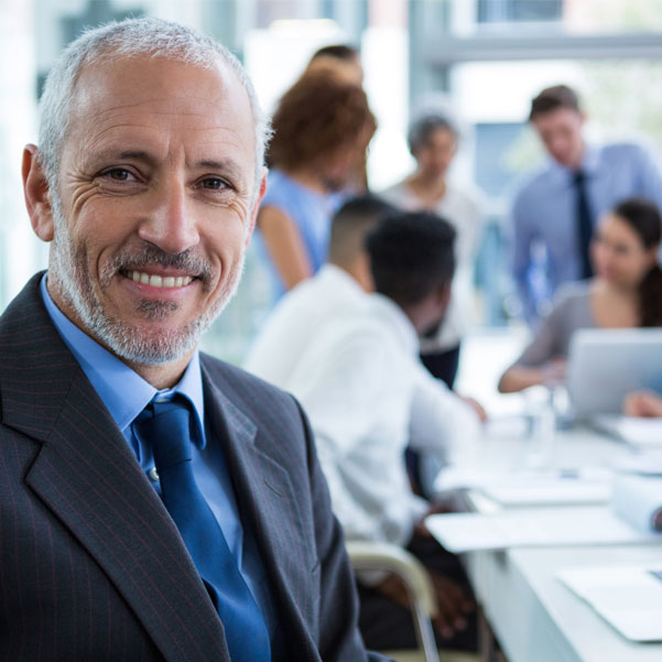 An older man in a business suit smiles to the camera. Behind him several people are having a meeting.
