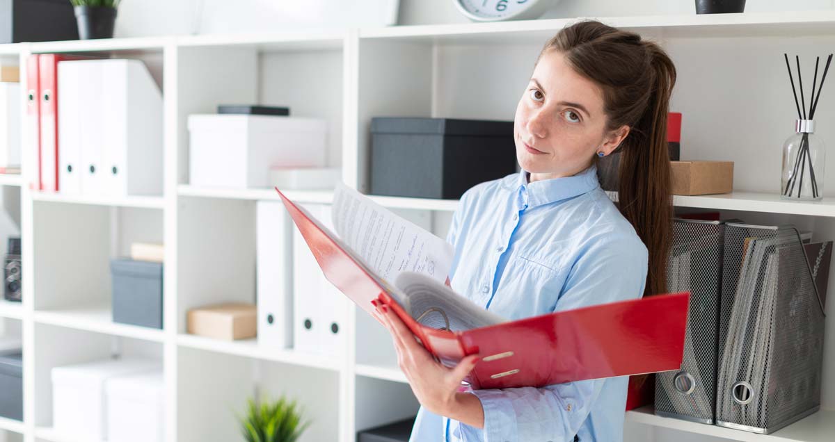 A young woman holding a thick level arch file looking serious and focused. A row of shelves with more files is behind her.