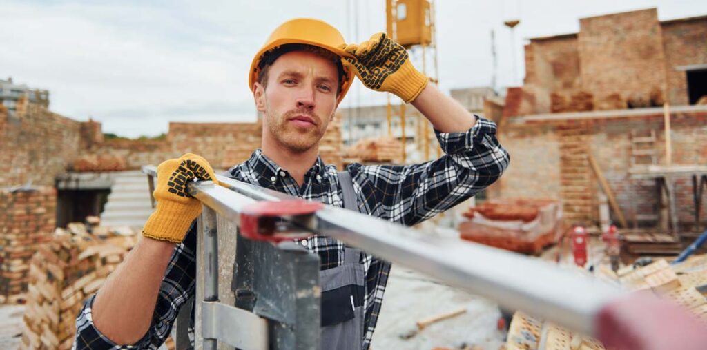 A white male construction worker in a yellow hard hat and gloves stands in front of a construction site, holding a ladder.