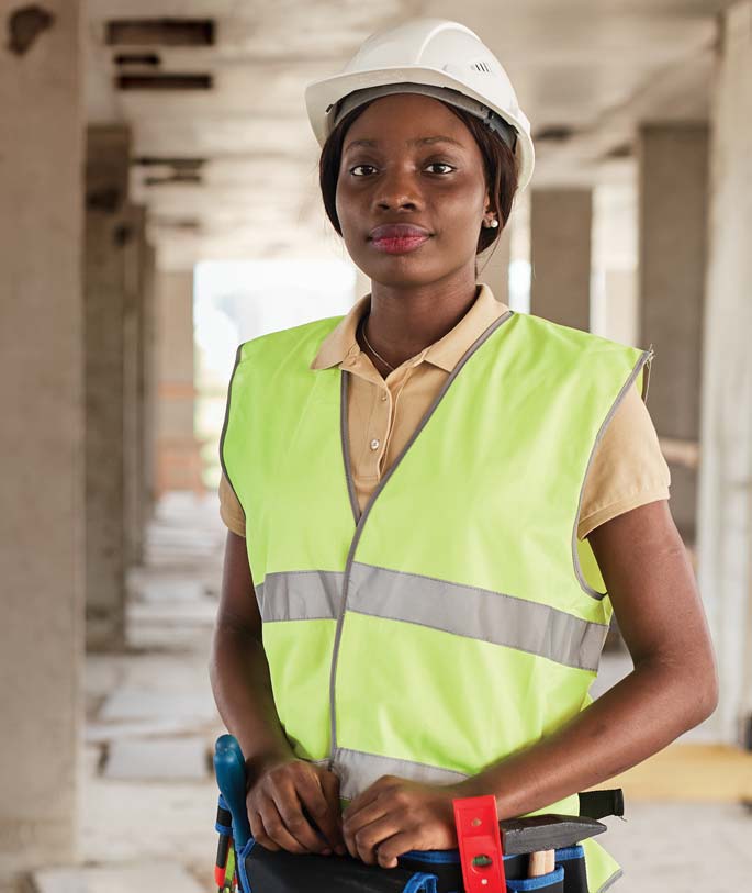 A black female construction worker wearing a white hard hat and a high-visibility safety vest on a construction site.