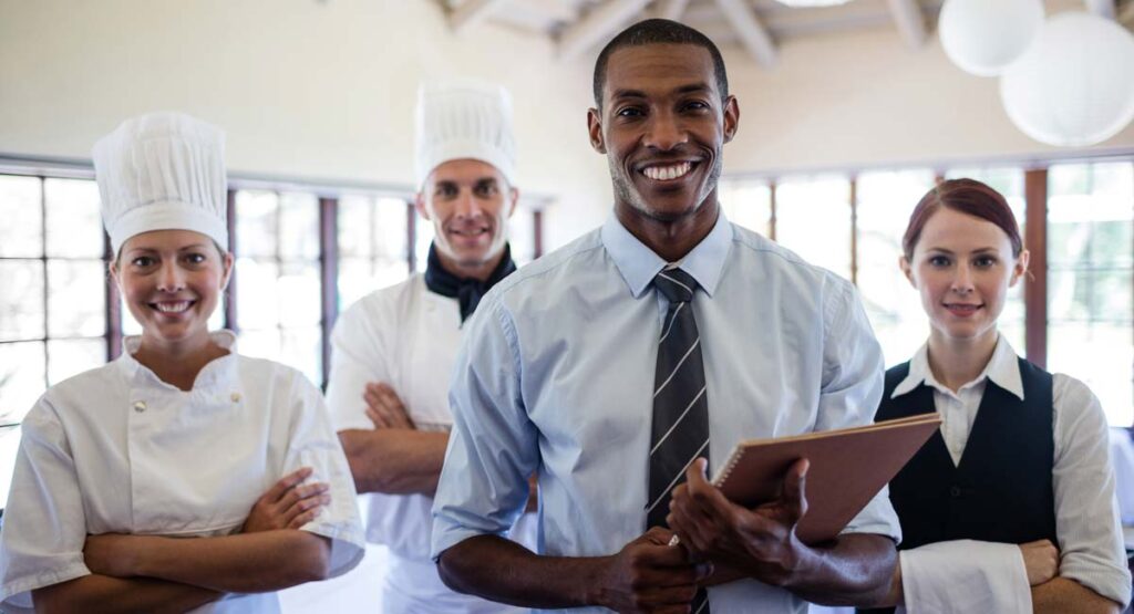 A diverse, smiling team of four restaurant staff members - two chefs, a manager with a clipboard, and a server - posing in a dining area.