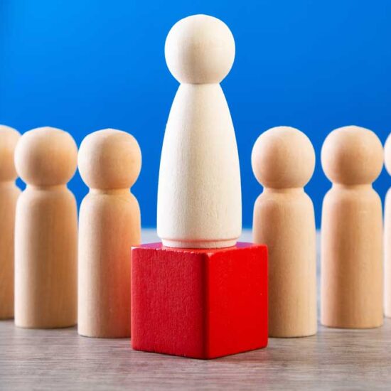 A large wooden peg doll elevated on a red cube in the centre of a line of six smaller peg dolls, set against a blue background.