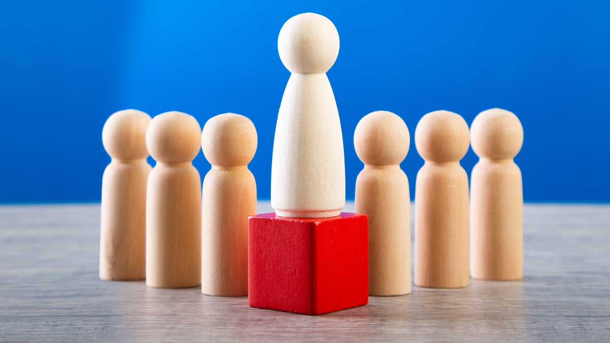 A large wooden peg doll elevated on a red cube in the centre of a line of six smaller peg dolls, set against a blue background.