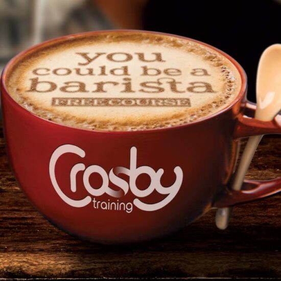 A red coffee cup filled with a latte on a wooden table. Text stencilled in the foam reads 'you could be a Barista'. The Crosby Training logo is on the side of the cup.