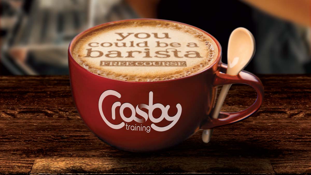 A red coffee cup filled with a latte on a wooden table. Text stencilled in the foam reads 'you could be a Barista'. The Crosby Training logo is on the side of the cup.