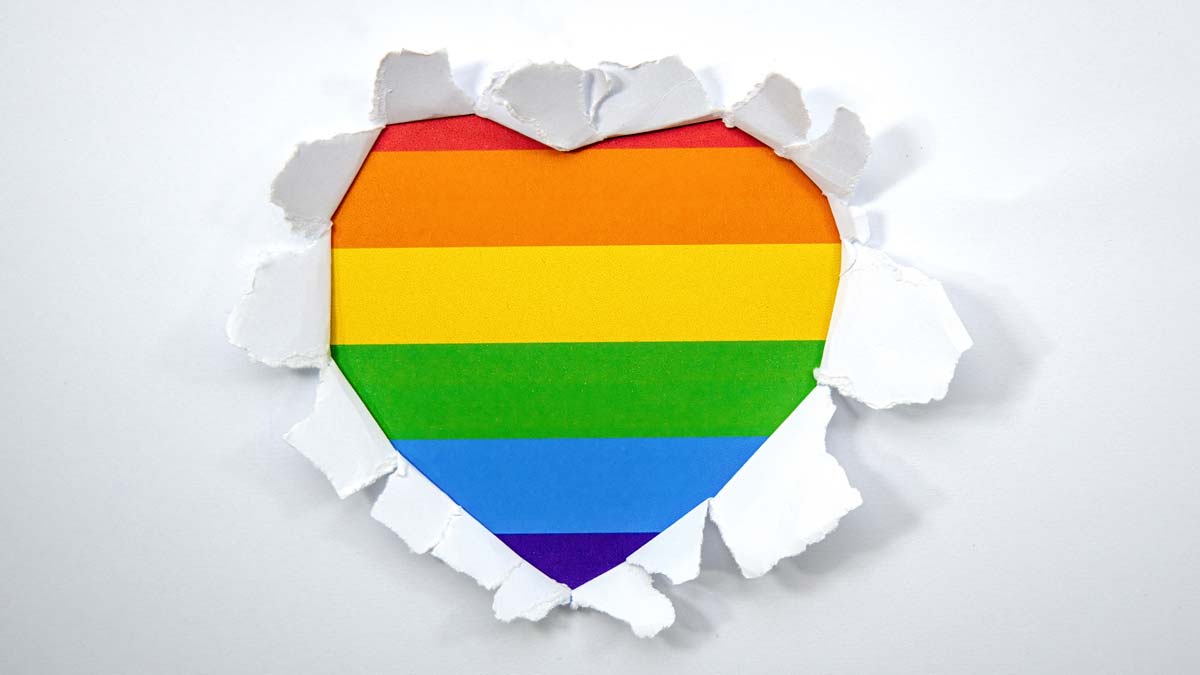 A heart-shaped rainbow Pride flag shown through torn white paper.
