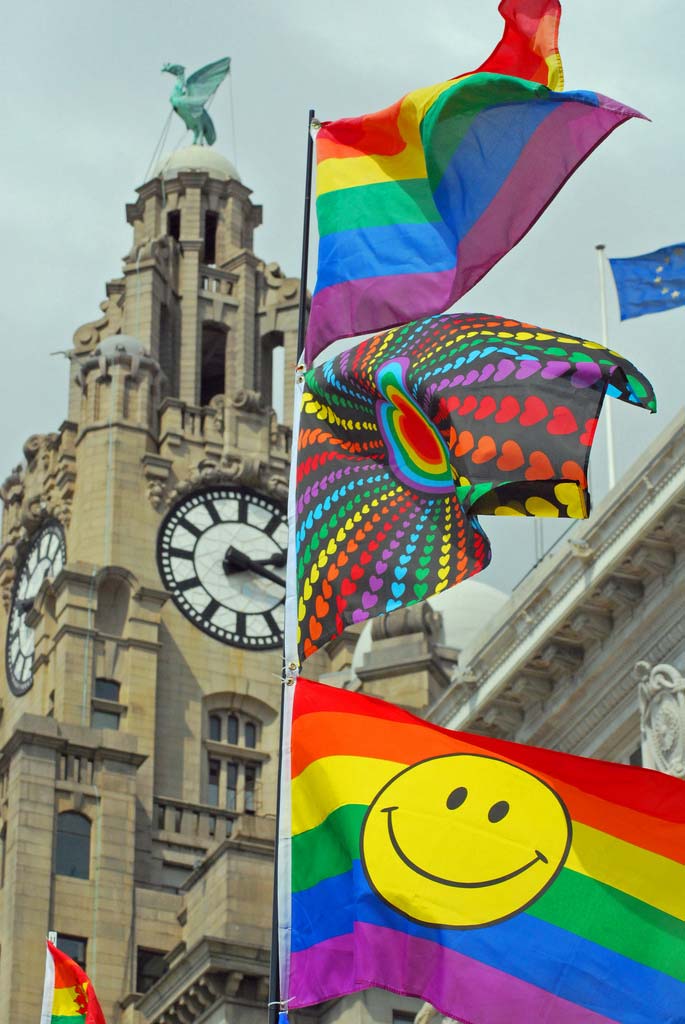 The iconic Liver Building. In the foreground is a collection of brightly coloured Pride flags featuring rainbow stripes, and one with a yellow smiley face.
