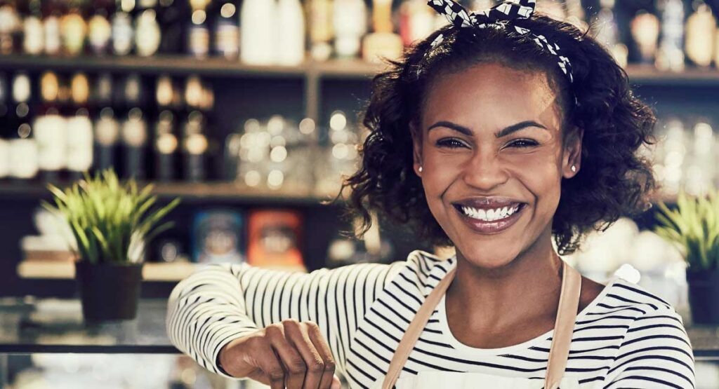 A smiling female barista wearing a striped shirt and light-coloured apron standing happily behind a coffee shop counter.
