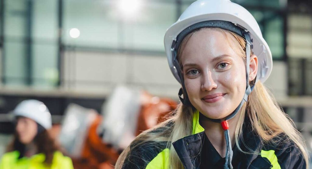 A young woman with blonde hair, wearing a white hard hat and a high-visibility jacket, smiles in an industrial setting.