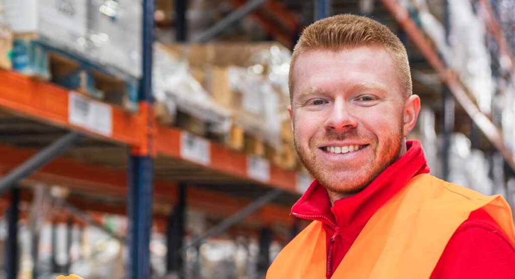A smiling man with red hair and a beard, wearing a red shirt and orange high-visibility vest, standing in a warehouse.