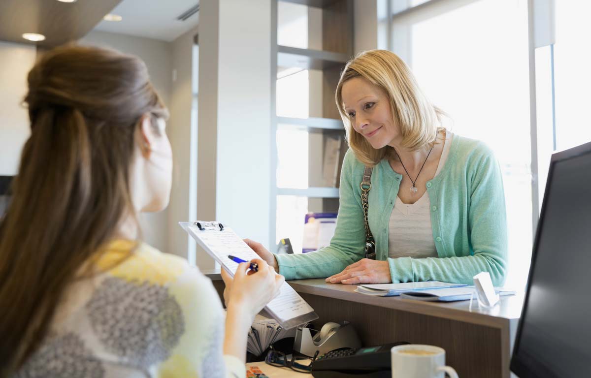 A middle aged woman at a reception desk being hand a form to fill in by the receptionist.