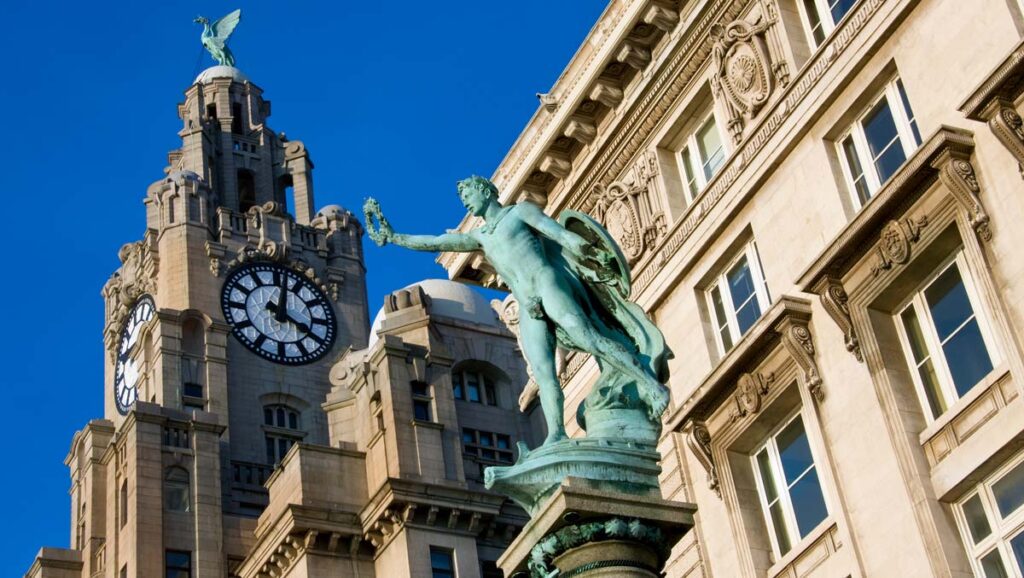 A bronze statue of a man in the foreground, with the ornate facade and clock tower of the historic Royal Liver Building in the background against a blue sky.