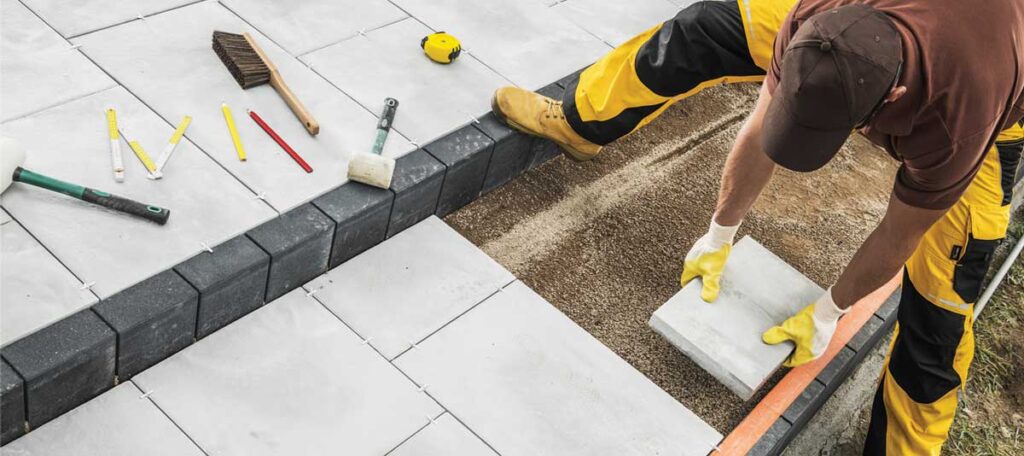 A man wearing yellow and black work clothes and gloves is installing a grey concrete paver into a base of sand next to an already laid section of pavers. Various tools are visible in the background.