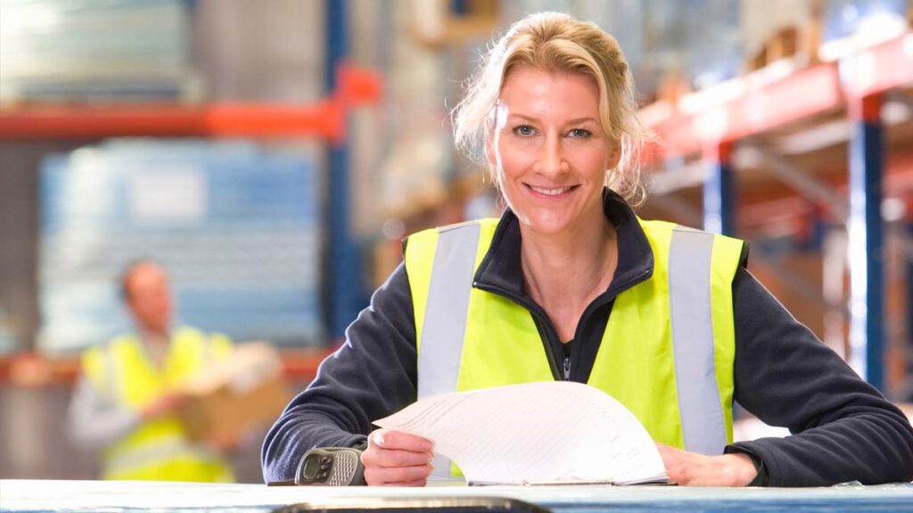 A smiling woman in a yellow high-visibility vest sits at a desk in a warehouse, holding papers.