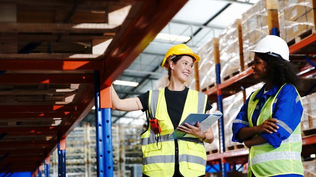 Two smiling female warehouse workers in hard hats and high-visibility vests stand talking in an aisle filled with industrial shelving and pallets.