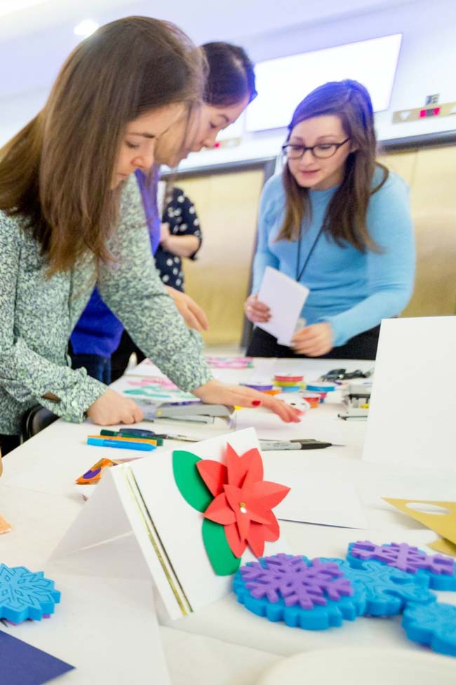 Three women standing around a white table doing a craft activity, with a handmade card featuring a red paper flower and blue and purple snowflake cutouts in the foreground.