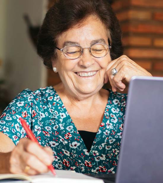 A smiling senior woman with dark hair and glasses, sitting at a table with a laptop open in front of her, using a pen to write in a notebook. 