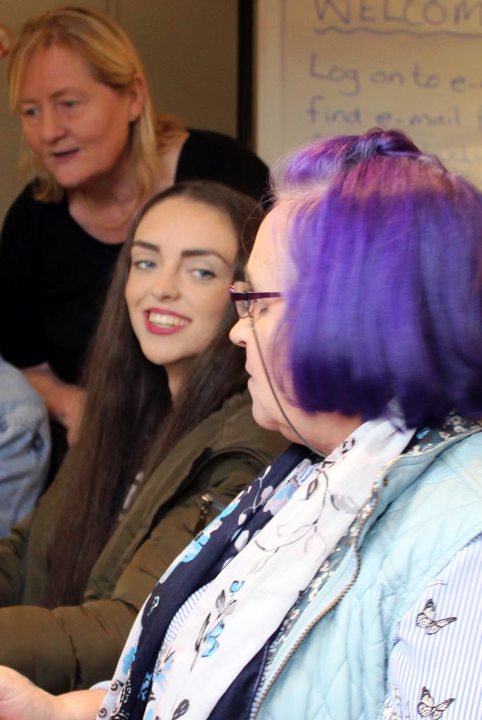 Two female learners using laptops: a young woman turns and grins broadly to an older woman with purple hair. A Crosby Training tutor stands behind them.