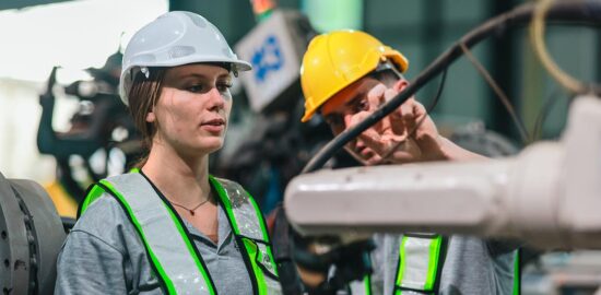 A female and a male engineer, wearing white and yellow hard hats and high-visibility vests, are working together in an industrial setting with machinery in the background.