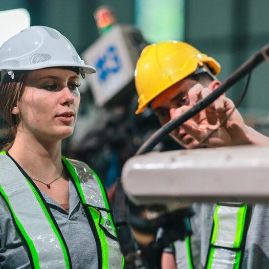 A female and a male engineer, wearing white and yellow hard hats and high-visibility vests, are working together in an industrial setting with machinery in the background.