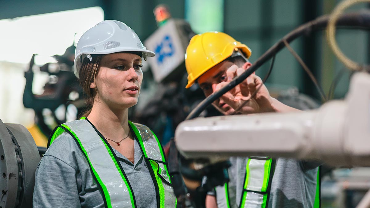 A female and a male engineer, wearing white and yellow hard hats and high-visibility vests, are working together in an industrial setting with machinery in the background.