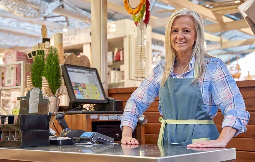 A smiling mature woman wearing a blue apron stands behind a store counter with a point-of-sale system in a clean, modern shopping area. 