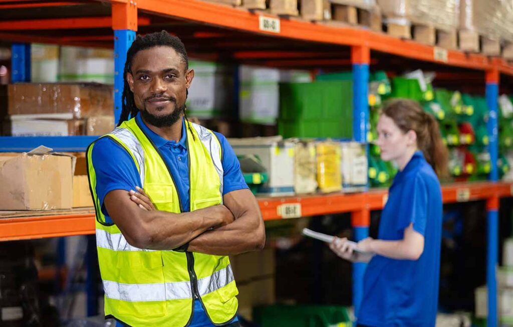 A confident male warehouse worker wearing a blue shirt and high-visibility safety vest stands with his arms crossed. In the background, a female colleague reviews inventory on a clipboard amidst warehouse shelving and stock. 