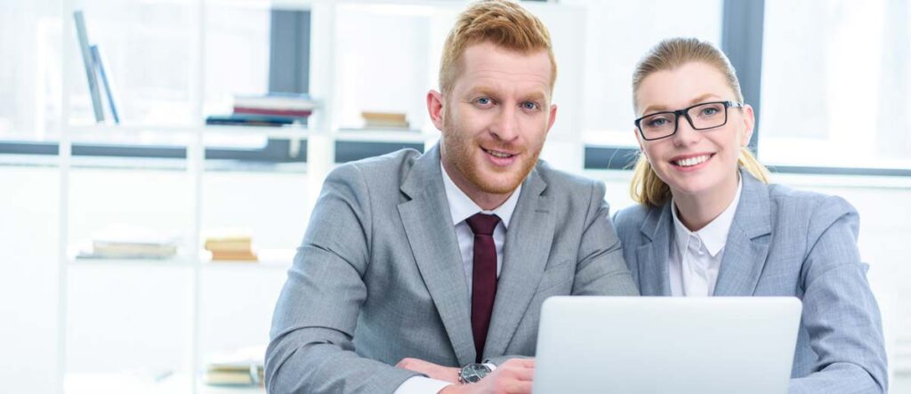 A man and a woman in business suits are sitting at a desk with a laptop smiling.