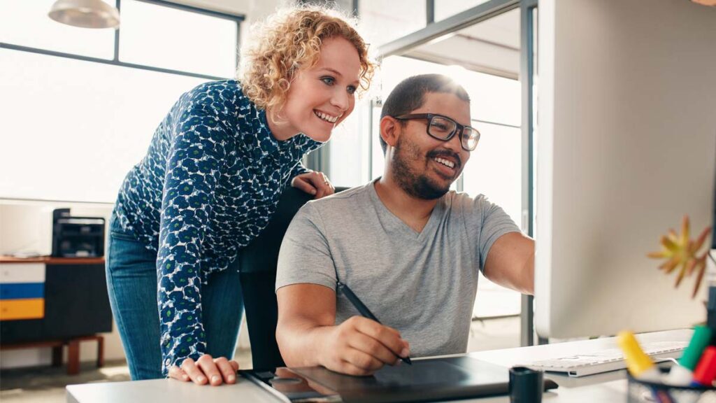 Two smiling colleagues collaborating on a design project, looking at a computer screen and using a drawing tablet with a stylus.