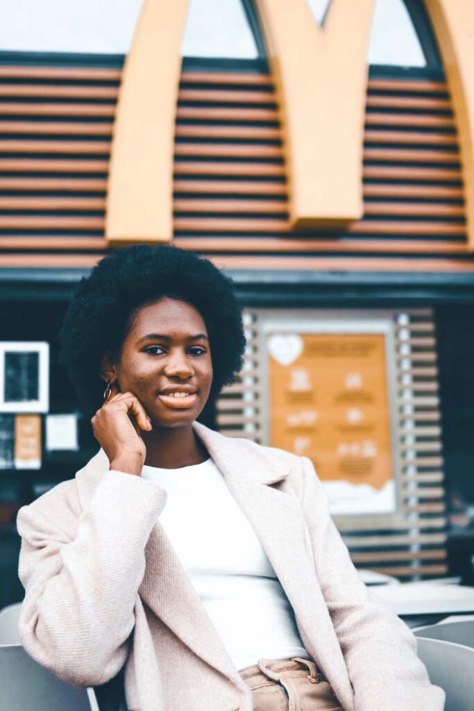 A young woman with an afro hairstyle, wearing a light beige coat,  sitting in front of a McDonald's restaurant.