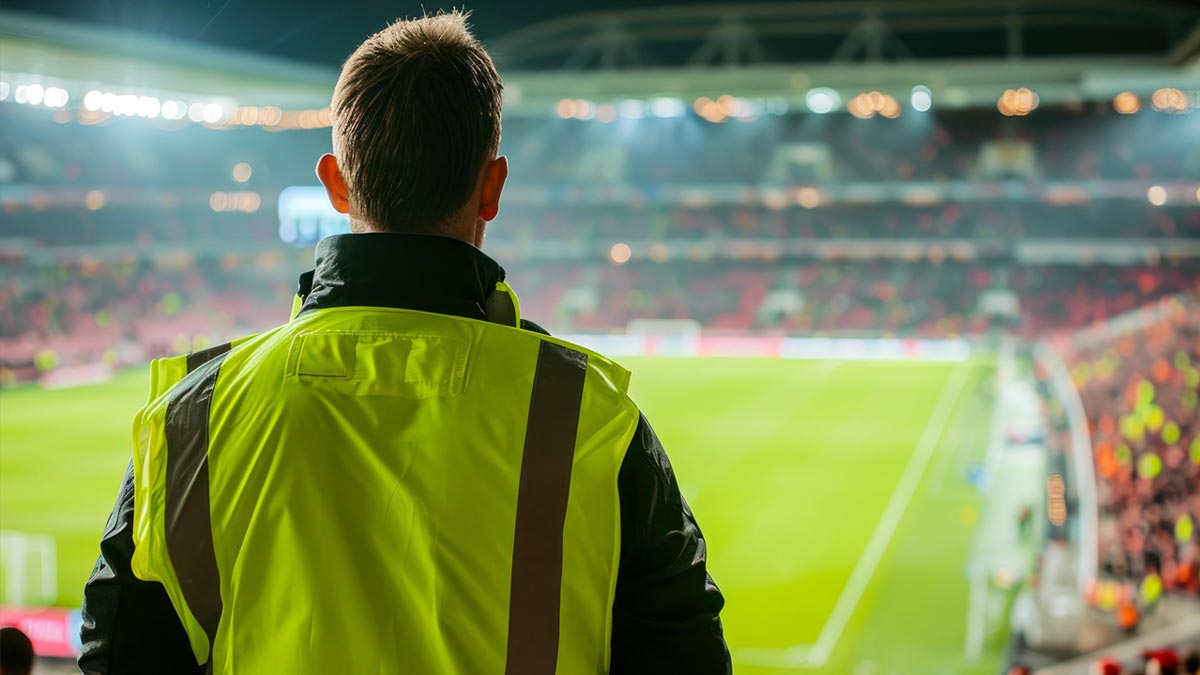 A staff member wearing a high-visibility yellow vest stands with their back to the camera, overlooking a crowded sports stadium pitch during a night event.