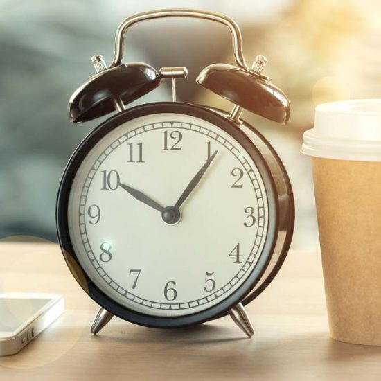An old fashioned alarm clock reads 5 past 10, there is a cup of coffee and a phone alongside it.